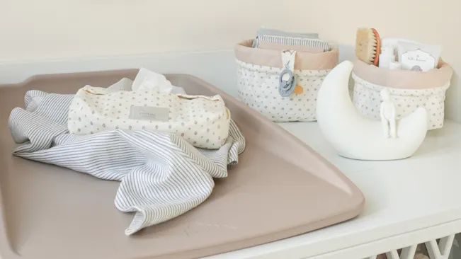 Pusleunderlag med stofble og vådservietter ligger på et puslebord, omgivet af små opbevaringskurve med babyplejeprodukter.