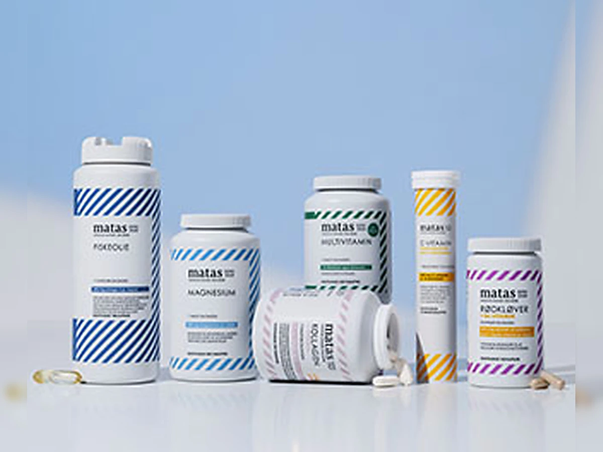 En hånd holder en stor bøtte Matas Striber fiskeolie ved siden af C-vitamin gummies og D-vitamin på lys baggrund.