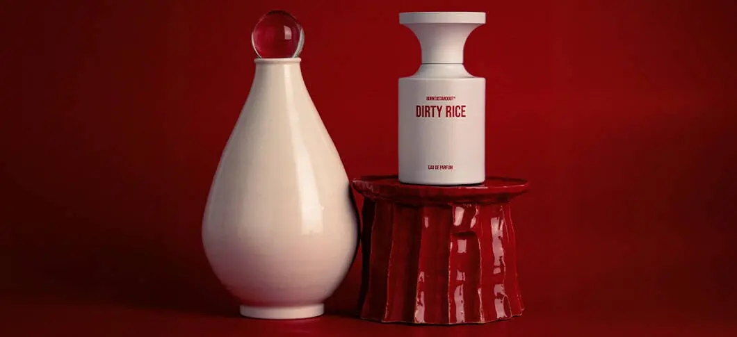 Her ses BORNTOSTANDOUT Dirty Rice Eau de Parfum i en hvid parfumeflakon med rød tekst, placeret på en blank rød sokkel. Ved siden af står en lys dekorativ vase, og billedet har en mørkerød baggrund.