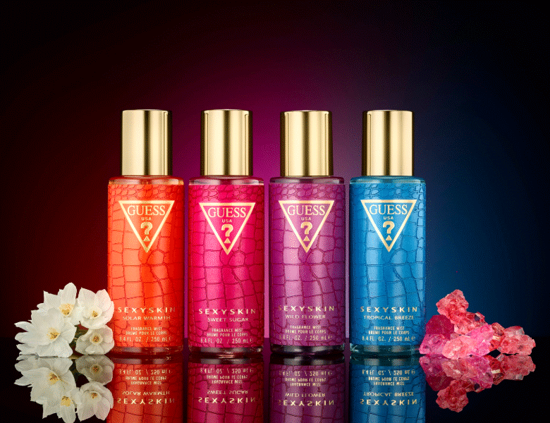 Her ses et billede med fire produkter fra GUESS placeret side om side mod en mørk baggrund med farvegradient. På produkterne står teksten “GUESS USA” og “SEXY SKIN” samt varianterne “SOLAR WARMTH”, “SWEET SUGAR”, “WILD FLOWER” og “TROPICAL BREEZE”. Produkterne er i orange, pink, lilla og blå nuancer med guldfarvede låg.