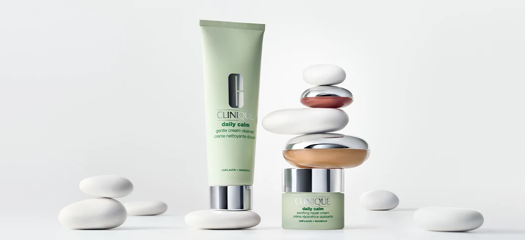 Her ses et billede med hudplejeprodukter fra Clinique placeret blandt glatte, hvide sten mod en lys baggrund. Produkterne omfatter “Daily Calm Gentle Cream Cleanser” og “Daily Calm Repair Cream”.