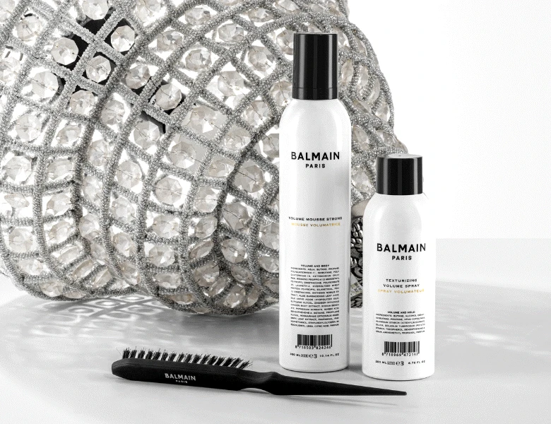 Her ses en hvid baggrund med en lysekrone. Foran ses en styling-hårbørste samt to hvide og sorte produkter.