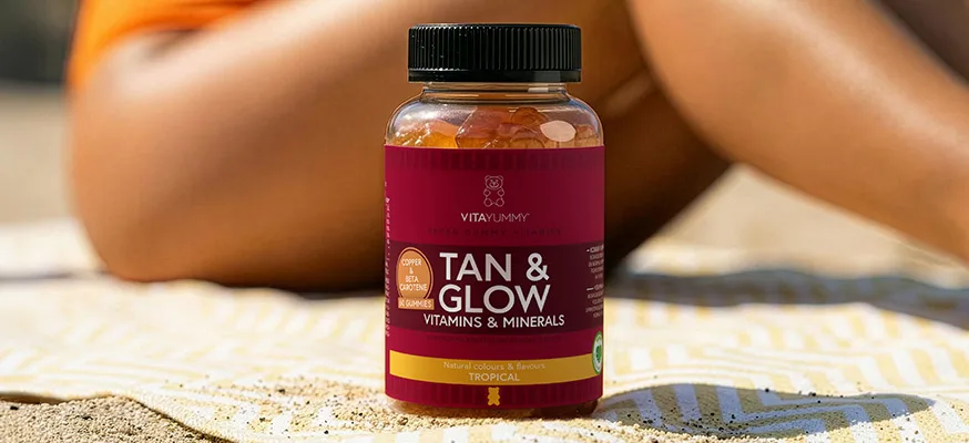 VitaYummy Tan & Glow gummies står på et håndklæde i sandet foran en solbadende person i orange badetøj.