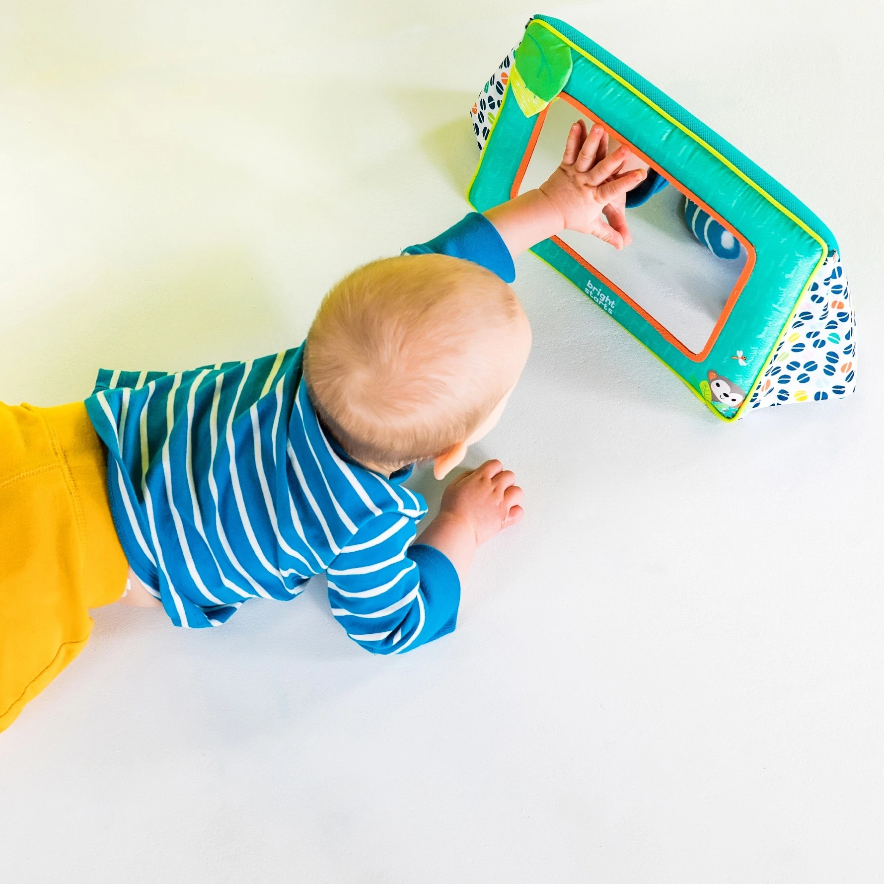 Baby ligger på maven og rækker ud efter et turkist Bright Starts spejllegetøj.