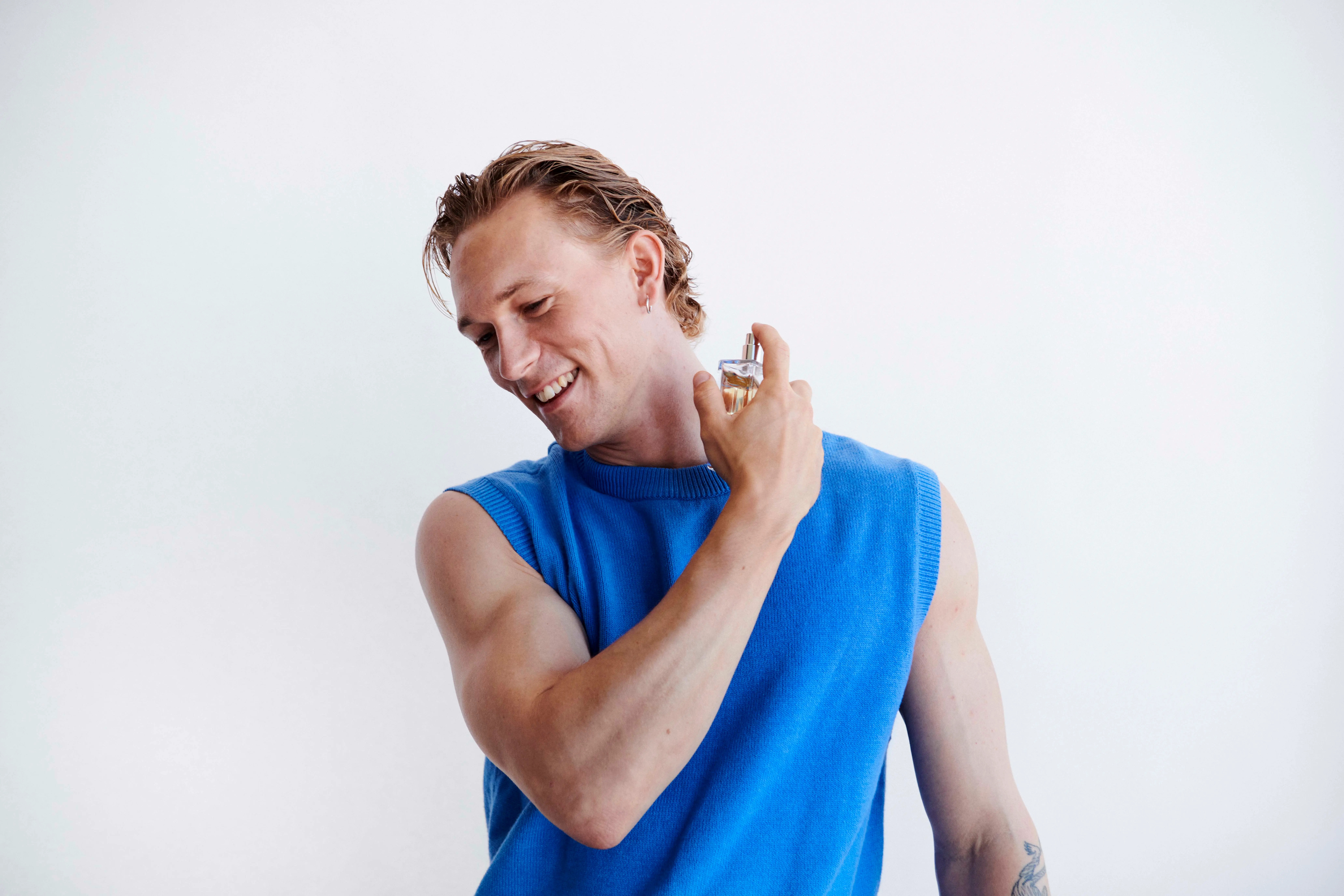 En smilende mand i en blå ærmeløs top, der sprayer parfume på sin hals.