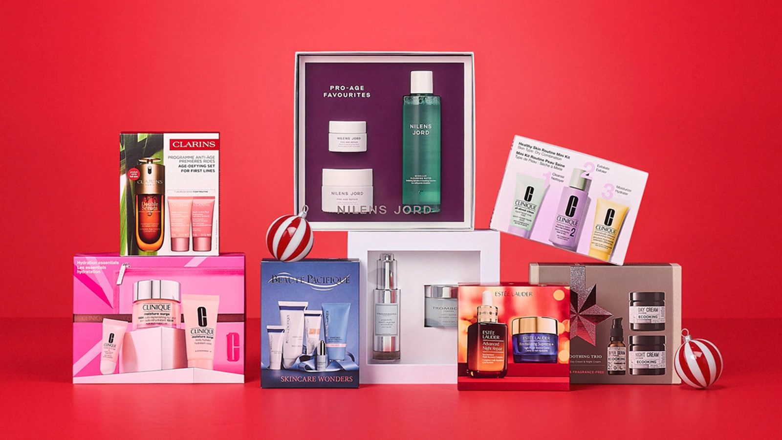 Hudpleje gaveæsker fra mærker som Clarins, Clinique, Estée Lauder og flere, arrangeret på rød baggrund.