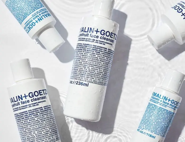 Her ses en hvid baggrund med fem hvide Malin+Goetz produkter til ansigtet. De har alle skrift i blå.