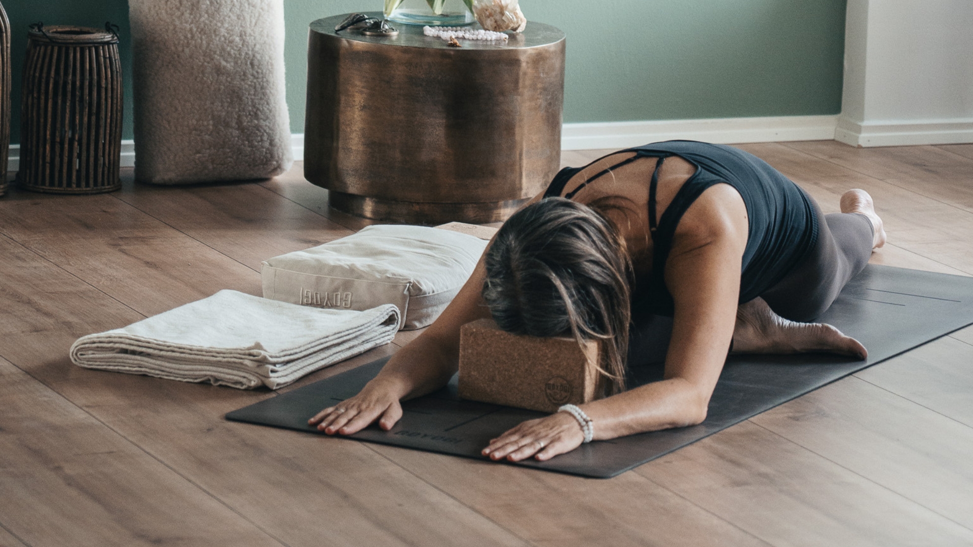 Kvinde laver en foroverbøjet yogaøvelse på en yogamåtte i et roligt hjemmemiljø med pude, tæppe og yogablok.