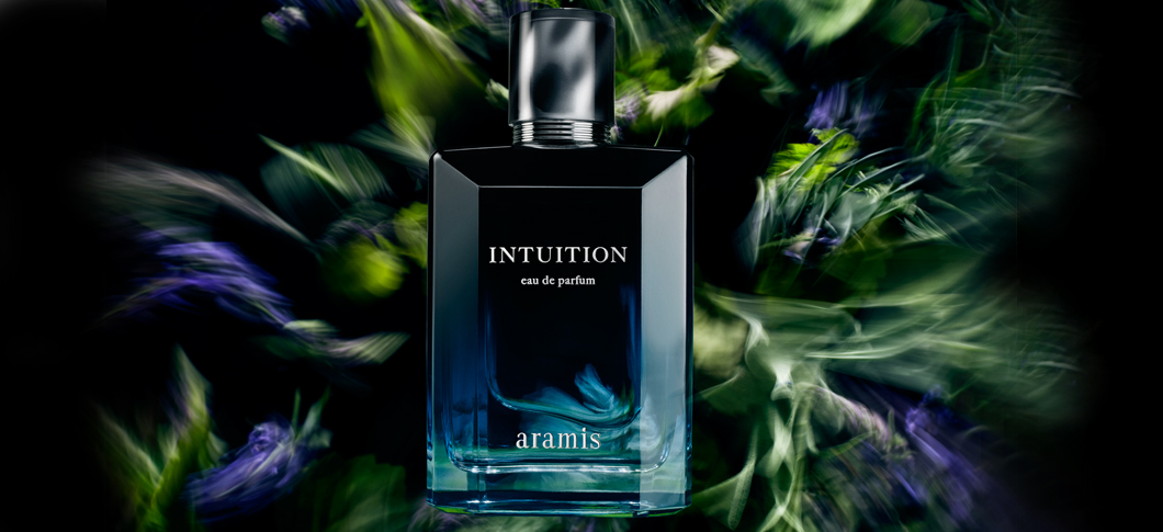 Her ses et billede af en parfumeflakon med teksten “INTUITION eau de parfum” og “aramis” placeret centralt mod en mørk baggrund med grønne og lilla bevægede planteelementer omkring.