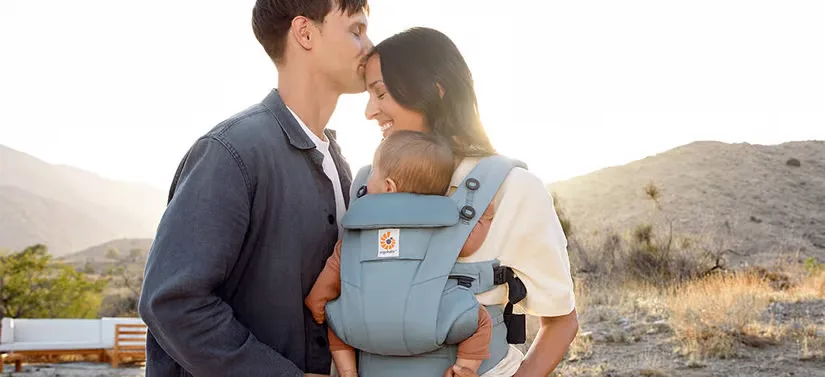 Her ses en mand, som kysser en kvinde på panden. Kvinden holder et barn i et Ergobaby produkt. I baggrunden er en smuk natur.