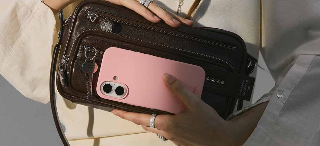 Her ses en hånd, der holder en mobil med lyserødt cover foran en brun taske.