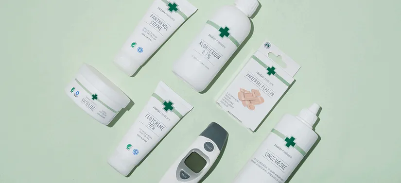 Fladt opstillet billede af forskellige medicinske og plejeprodukter på en lys grøn baggrund. Produkterne inkluderer blandt andet creme, vaseline, håndsprit, plaster, linsevæske og et digitalt termometer. Emballagen er hvid med grønne kors og grøn tekst, hvilket giver et rent og klinisk udtryk.
