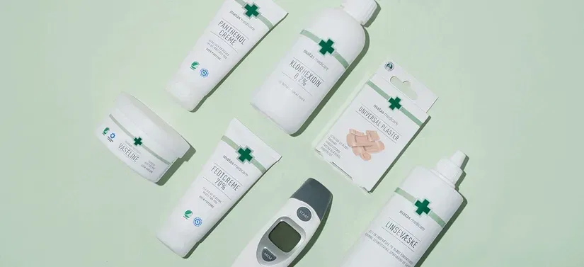 Fladt opstillet billede af forskellige medicinske og plejeprodukter på en lys grøn baggrund. Produkterne inkluderer blandt andet creme, vaseline, håndsprit, plaster, linsevæske og et digitalt termometer. Emballagen er hvid med grønne kors og grøn tekst, hvilket giver et rent og klinisk udtryk.