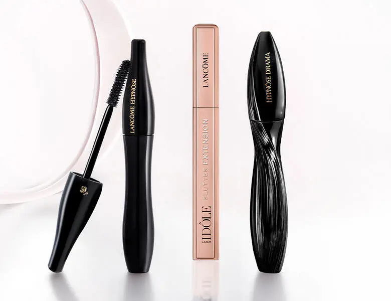 Her ses et billede med tre mascaraprodukter fra Lancôme placeret på en lys, reflekterende overflade mod en lys baggrund. På produkterne står teksten “LANCÔME HYPNÔSE”, “IDÔLE FLUTTER EXTENSION” og “HYPNÔSE DRAMA”. Den ene mascara er vist åben med børsten synlig.