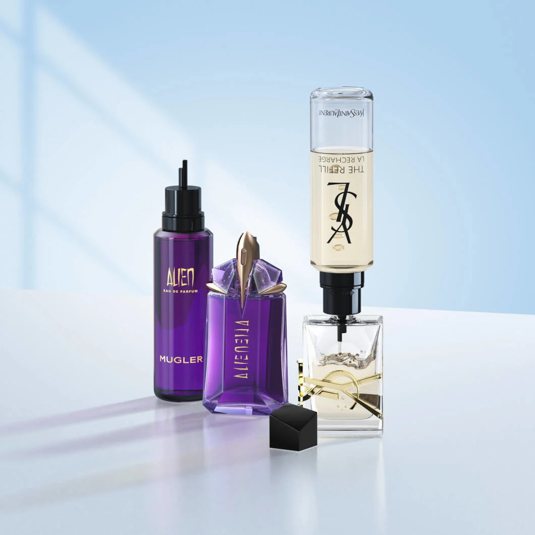 Tre parfumeprodukter står på en lys overflade foran en blød lyseblå baggrund. Til venstre ses en lilla refillflaske fra Mugler med teksten “Alien Eau de Parfum”. I midten står den ikoniske lilla Alien-parfumeflaske med kantet design og gulddetaljer. Til højre ses en Yves Saint Laurent-parfume, hvor en refillbeholder er monteret oven på parfumeflasken for at vise genopfyldning. Produkterne er opstillet i et stilrent og moderne miljø med fokus på luksusparfumer og refill-løsninger.