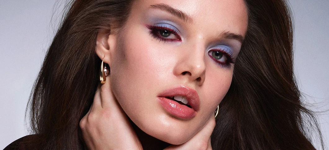 Her ses et billede af en person med makeup mod en lys baggrund. Personen har lyseblå øjenskygge, rosa læber og guldfarvede øreringe og ser direkte mod kameraet.