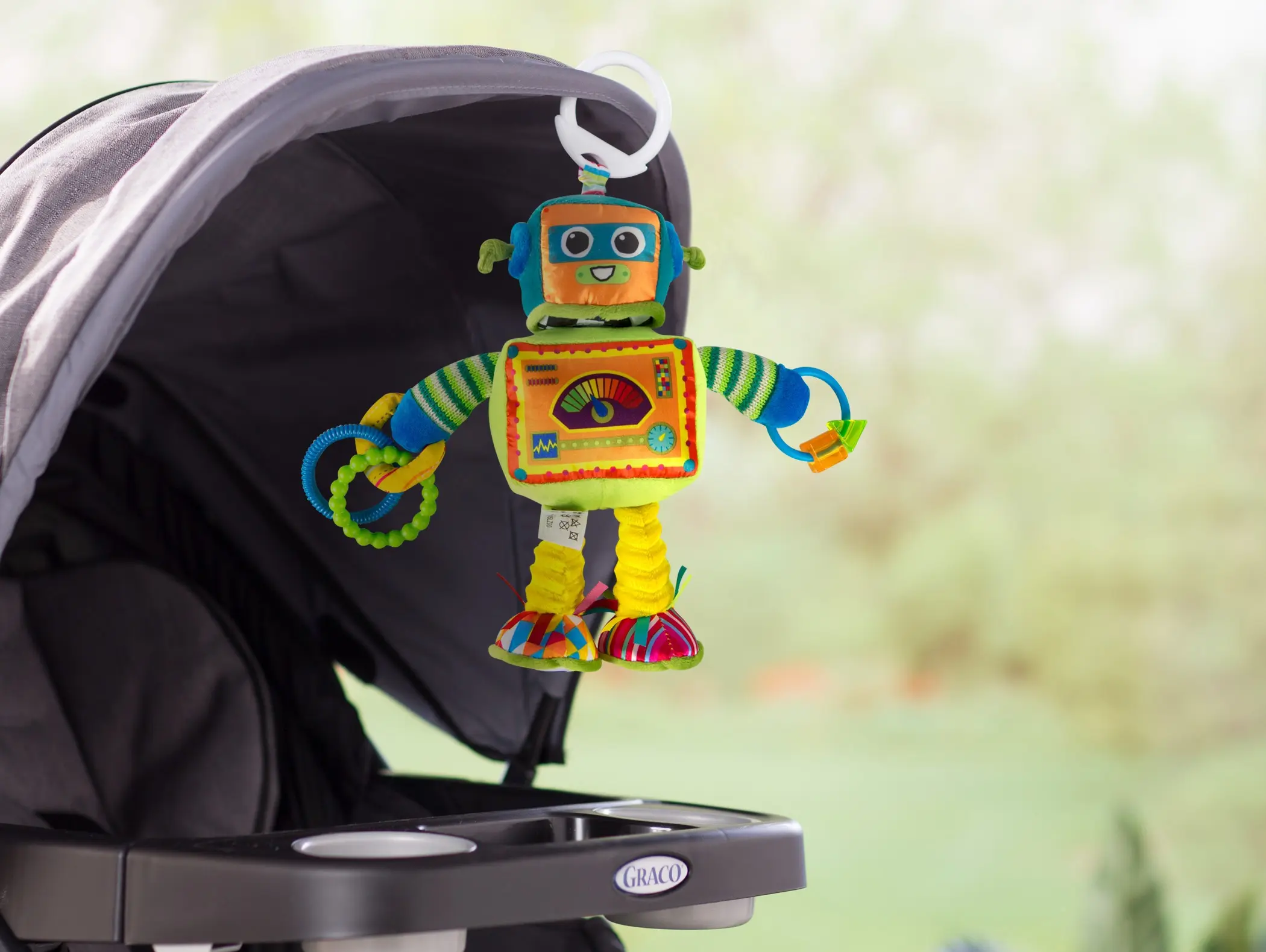 Farverigt robot-formet aktivitetslegetøj med striber og ringe, der hænger fra kalechen på en mørkegrå Graco klapvogn.