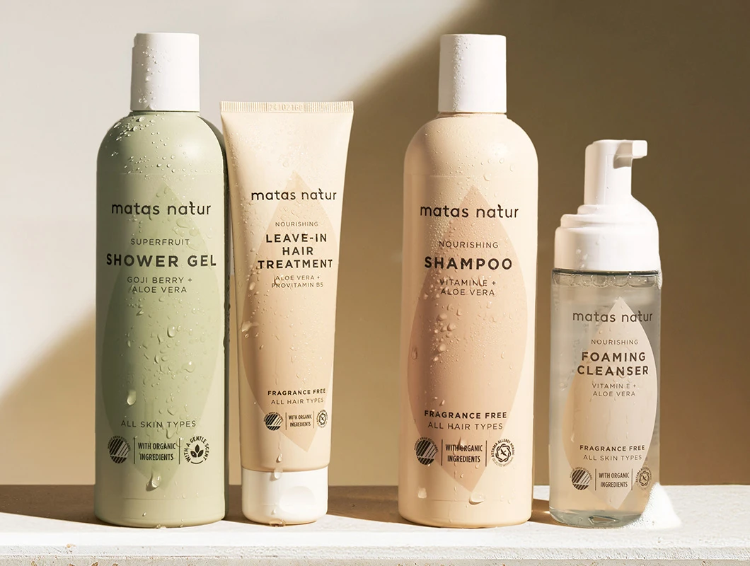 Her ses et billede med fire produkter fra Matas Natur mod en varm, lys baggrund. Produkterne omfatter Superfruit Shower Gel, Nourishing Leave-In Hair Treatment, Nourishing Shampoo og Nourishing Foaming Cleanser. Emballagen er dækket af små vanddråber, og produkterne er placeret på en lys overflade med blødt sollys, der skaber et naturligt og friskt udtryk.
