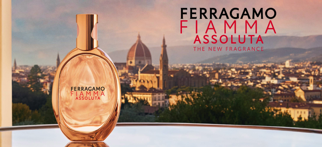 Her ses et billede af en parfumeflakon fra Ferragamo placeret foran en byudsigt ved solnedgang. Øverst til højre står teksten “FERRAGAMO FIAMMA ASSOLUTA THE NEW FRAGRANCE”. Flakonen er i varme rosagyldne nuancer med guldfarvet låg.