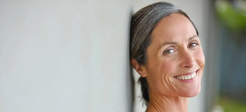 Nærbillede af en smilende kvinde med gråt hår, der læner sig op ad en lys væg.