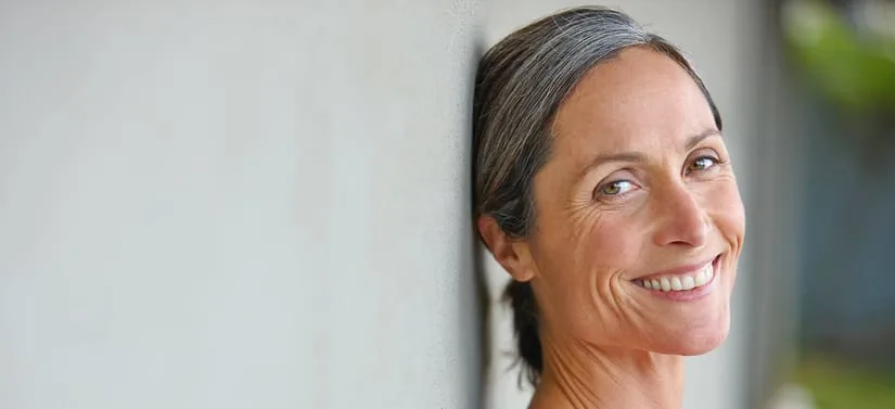 Nærbillede af en smilende kvinde med gråt hår, der læner sig op ad en lys væg.