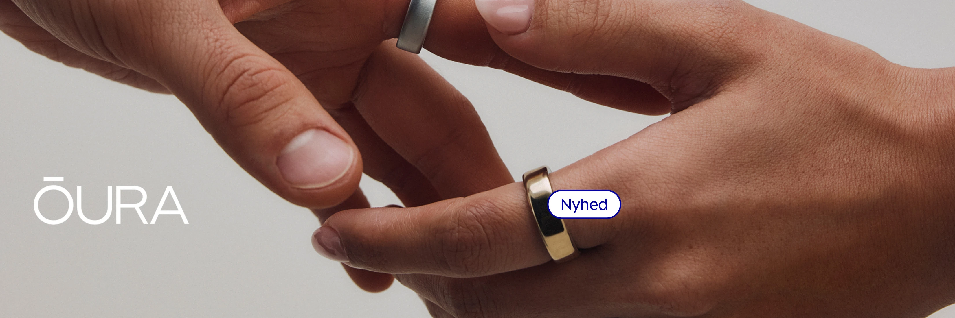 Nærbillede af to hænder, den ene med en sølvfarvet OURA-ring og den anden med en guldfarvet OURA-ring. Tekst: Nyhed'.