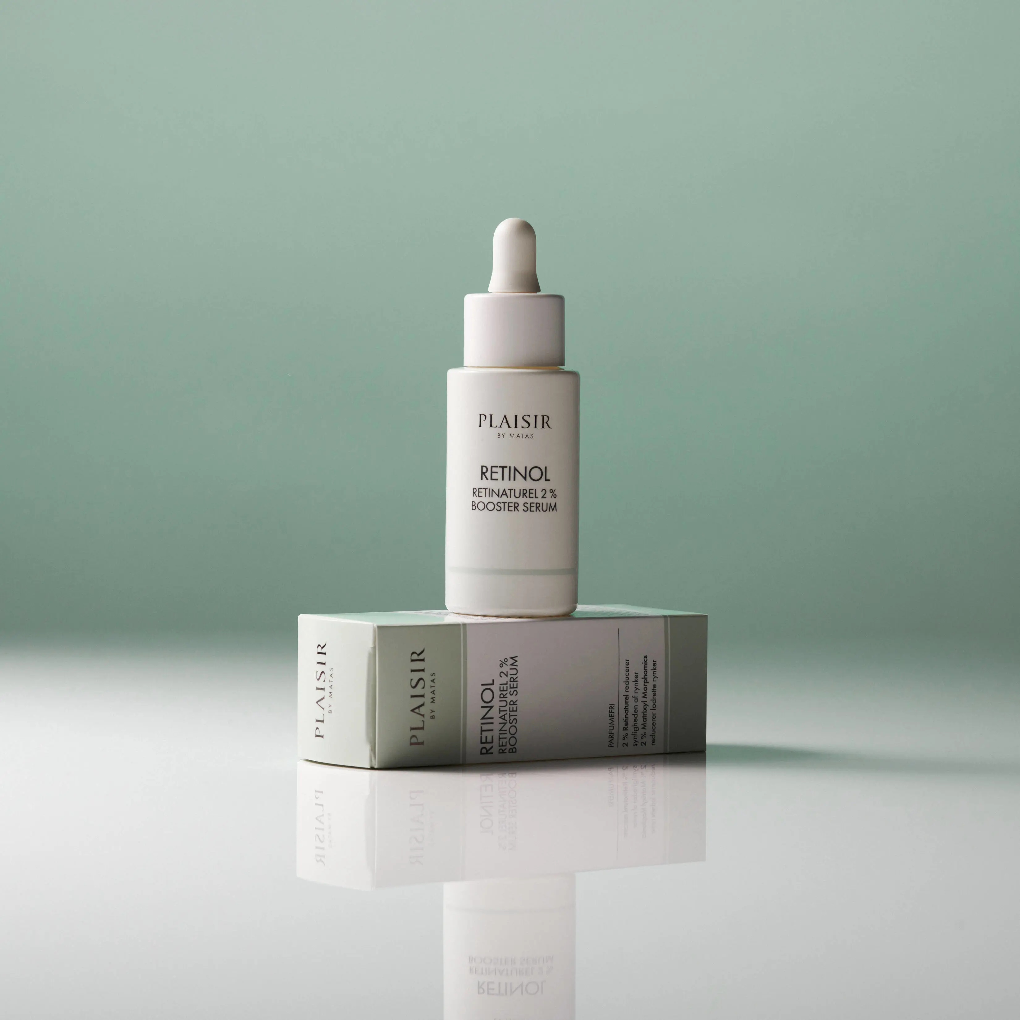En flaske Plaisir Retinol 2% Booster Serum står oven på sin æske på en blank overflade mod en grøn baggrund. Flasken har pipette og spejler sig i underlaget.