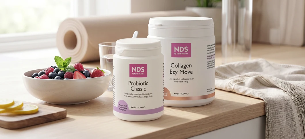 To NDS kosttilskud står på et træbord i et lyst køkkenmiljø ved siden af en skål med bær, et glas vand, en yogamåtte og et lyst viskestykke.