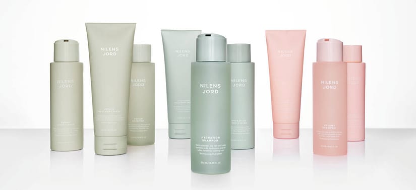 Her ses et billede med flere hårplejeprodukter fra Nilens Jord placeret side om side mod en lys, reflekterende baggrund. På produkterne står teksten “NILENS JORD” samt varianter som “Repair Shampoo”, “Hydration Shampoo” og “Volume Shampoo”. Produkterne er i lyse grønne og rosa nuancer.