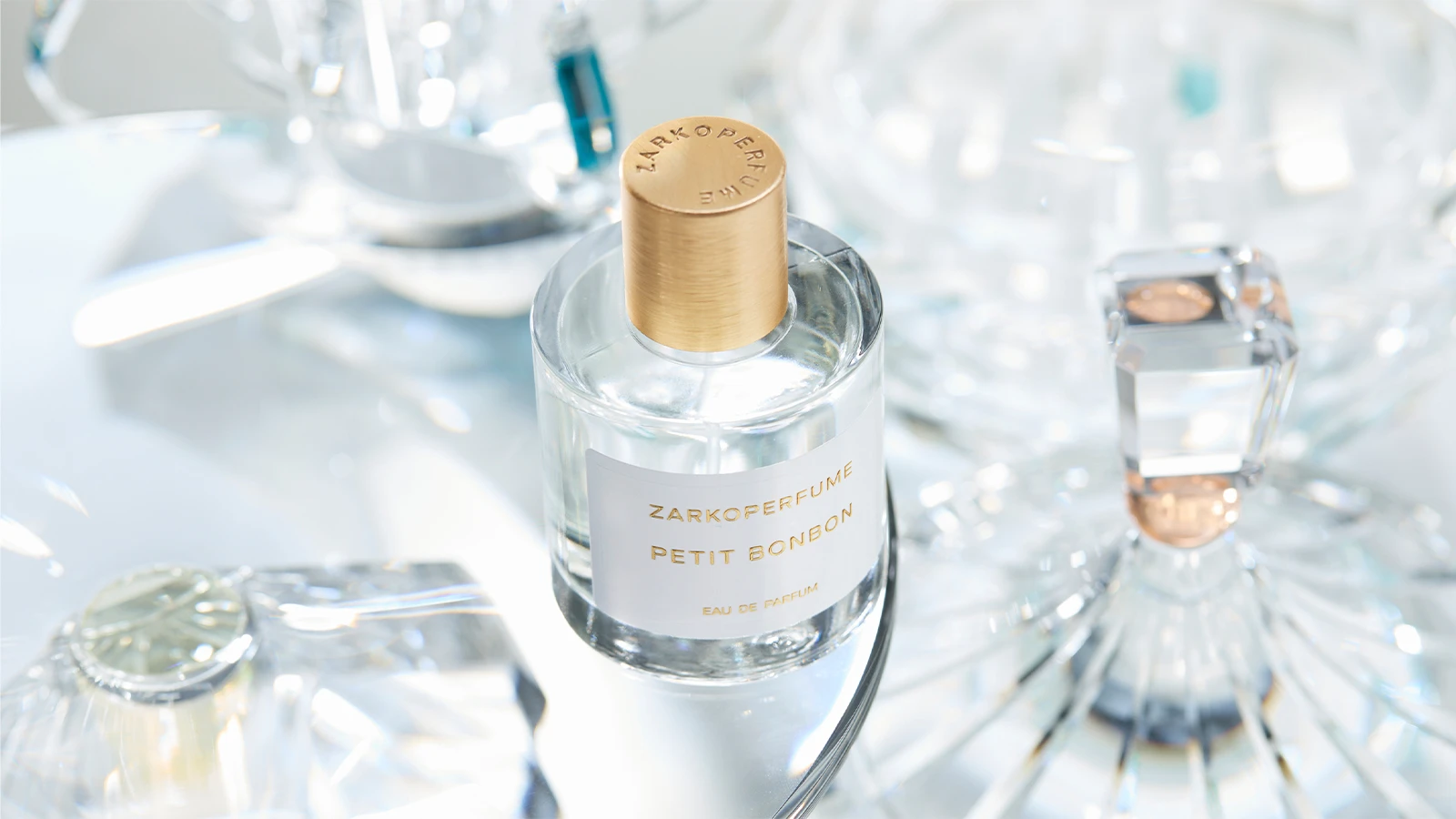 Her ses Zarkoperfume Petit Bonbon Eau de Parfum i en transparent parfumeflakon med guldfarvet låg og hvid etiket. Flakonen står blandt transparente glasdetaljer, der reflekterer lyset.