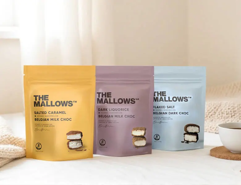 Gul, lilla og lyseblå The Mallows poser i et lyst miljø med en hvid kop med te til højre og et lyst tæppe i baggrunden. De står på et lyst bord