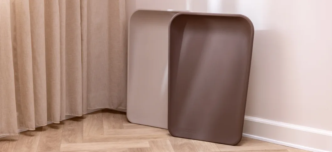 Her ses to Leander opbevaringsbakker i beige og brun placeret på et trægulv ved et gardin.