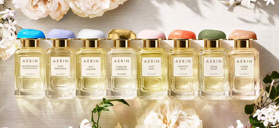 Her ses otte AERIN-parfumeflakoner med guldfarvede detaljer og låg i forskellige farver. Duftene står på række omgivet af lyse blomster.