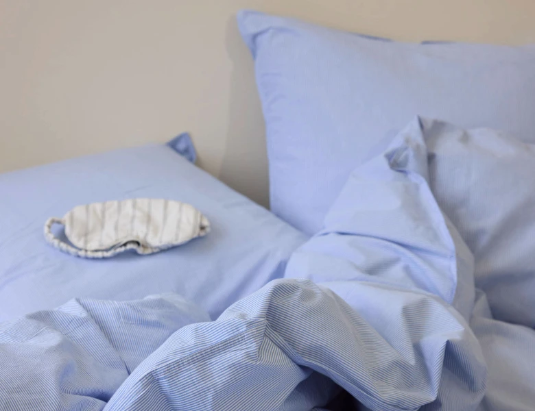 Her ses lyseblåt stribet sengetøj med puder og en sovemaske placeret på sengen.