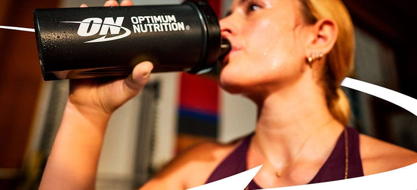 Her ses nogle en kvinde fra siden, som drikker af en sort Optimum Nutrition drikkedunk. Det ligner der et en sløret træningsmaskine bag hende.