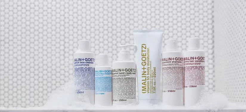 Her ses en hvid baggrund med noget sæbeskum. I midten ses seks hvide Malin+Goetz produkter med skrift i forskellige farver.