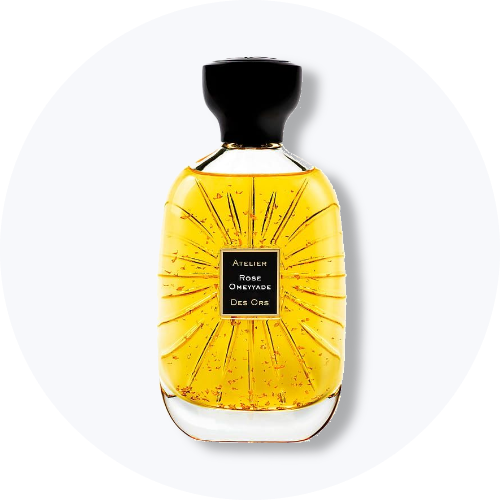 Her ses et billede af en parfumeflakon fra Atelier Des Ors placeret centralt mod en lys, rund baggrund. På flakonen står teksten “ROSE OMEYYADE DES ORS”. Flakonen er i gyldne nuancer med sort låg og dekorative strålemønstre på glasset.