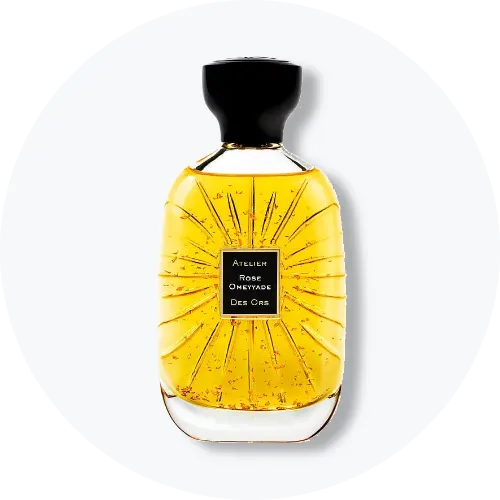 Her ses et billede af en parfumeflakon fra Atelier Des Ors placeret centralt mod en lys, rund baggrund. På flakonen står teksten “ROSE OMEYYADE DES ORS”. Flakonen er i gyldne nuancer med sort låg og dekorative strålemønstre på glasset.