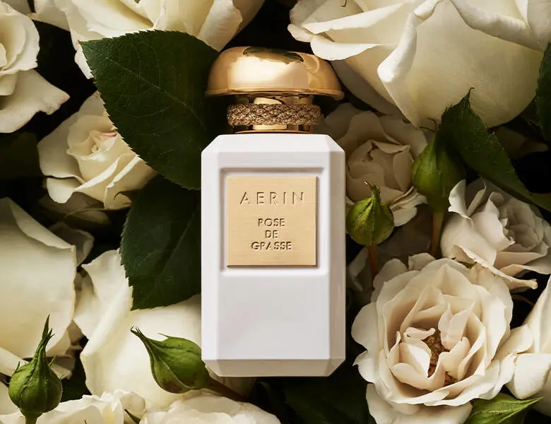 Her ses AERIN Rose de Grasse i en hvid parfumeflakon med guldfarvet låg og etiket, omgivet af hvide roser og grønne blade.