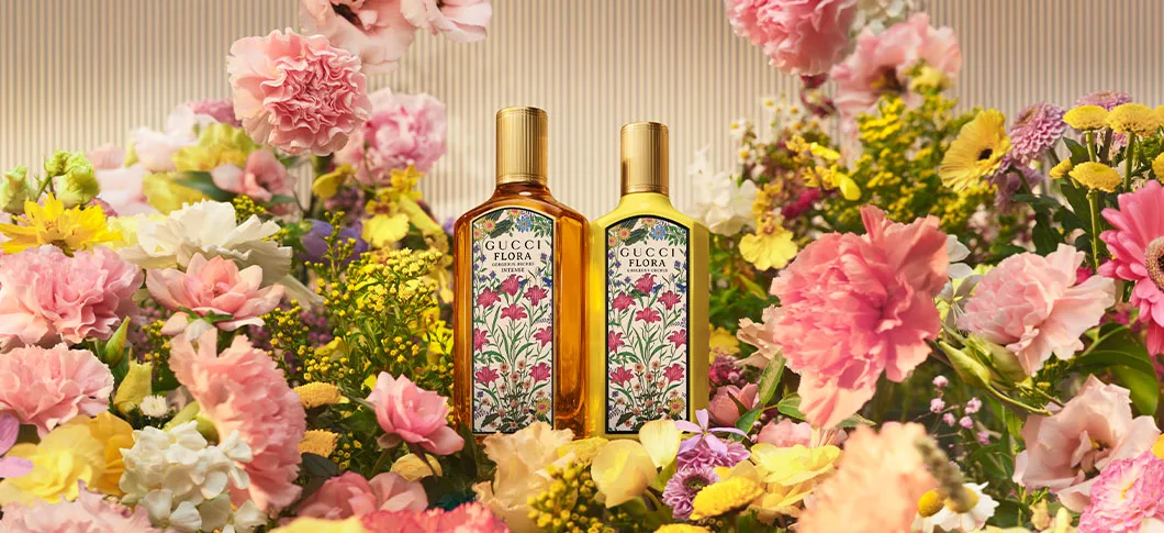 Her ses et billede med to parfumeflakoner fra Gucci Flora placeret blandt farverige blomster mod en lys baggrund. Flakonerne har blomstrede etiketter og gyldne låg og er omgivet af lyserøde, gule og hvide blomster.