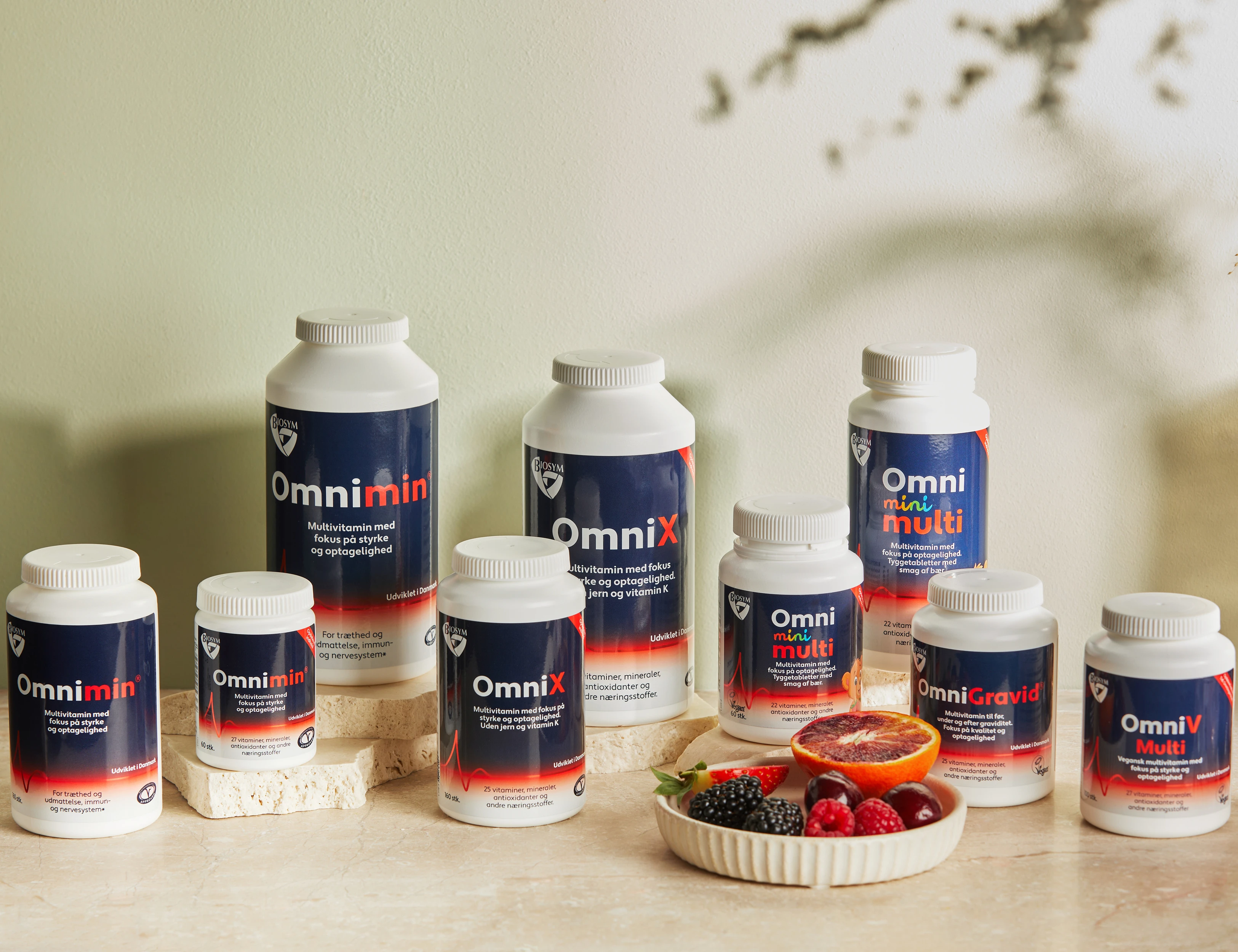 Flere Biosym multivitaminer står samlet på lyse stenplader foran en lys grøn baggrund med en skål fyldt med bær og en halveret blodappelsin.