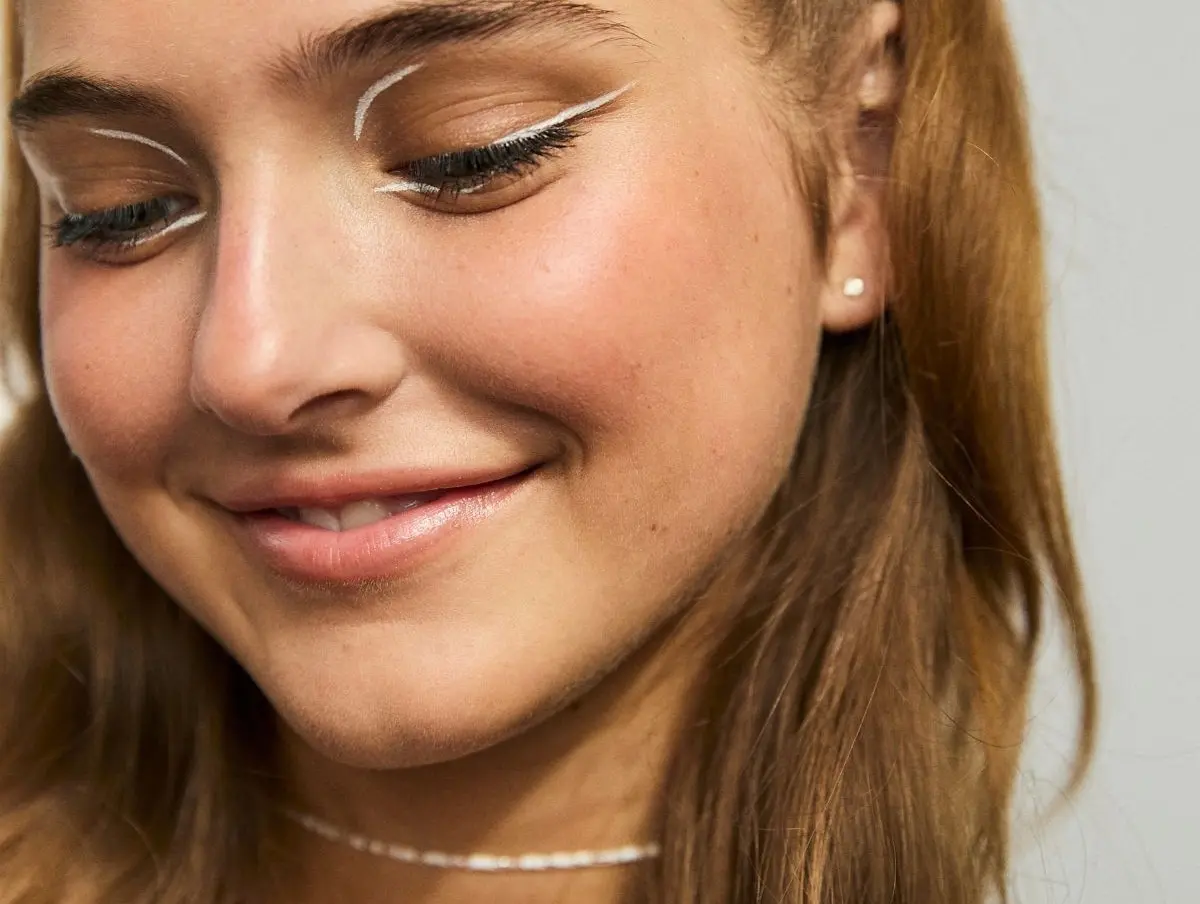 Nærbillede af en smilende person med hvid grafisk eyeliner, blød rouge og en hvid perlekæde.