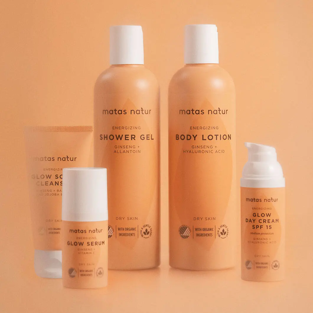 Flere Matas Natur Energizing hud- og kropsplejeprodukter står samlet mod en varm orange baggrund. Produkterne inkluderer shower gel, body lotion, glow serum, glow scrub cleanser og dagcreme med pumpe. Emballagen er i matchende orange nuancer.