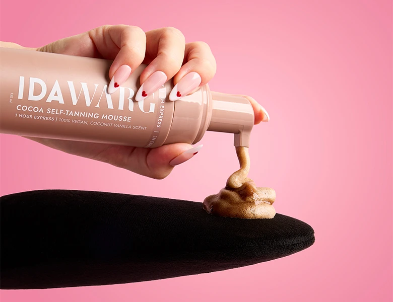 Her ses en hånd, der påfører IDA WARG selvbruner mousse på en handske foran en lyserød baggrund.