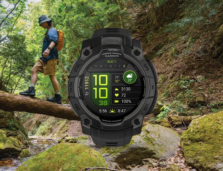 Person vandrer i skoven og balancerer på en træstamme over en bæk, mens et robust Garmin smartwatch med aktivitetsdata vises stort i forgrunden.