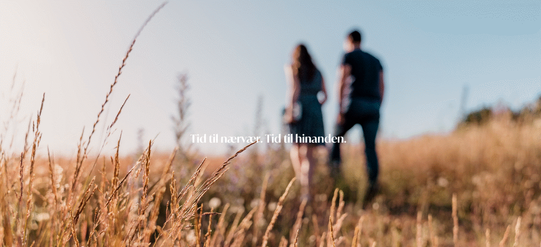 Et varmt, solbelyst landskab med højt, gyldent græs i forgrunden. I baggrunden går en mand og en kvinde side om side væk fra kameraet, let ude af fokus. Stemningen er rolig og sommerlig. Midt i billedet står teksten: “Tid til nærvær. Tid til hinanden.” i en lys, diskret skrifttype.