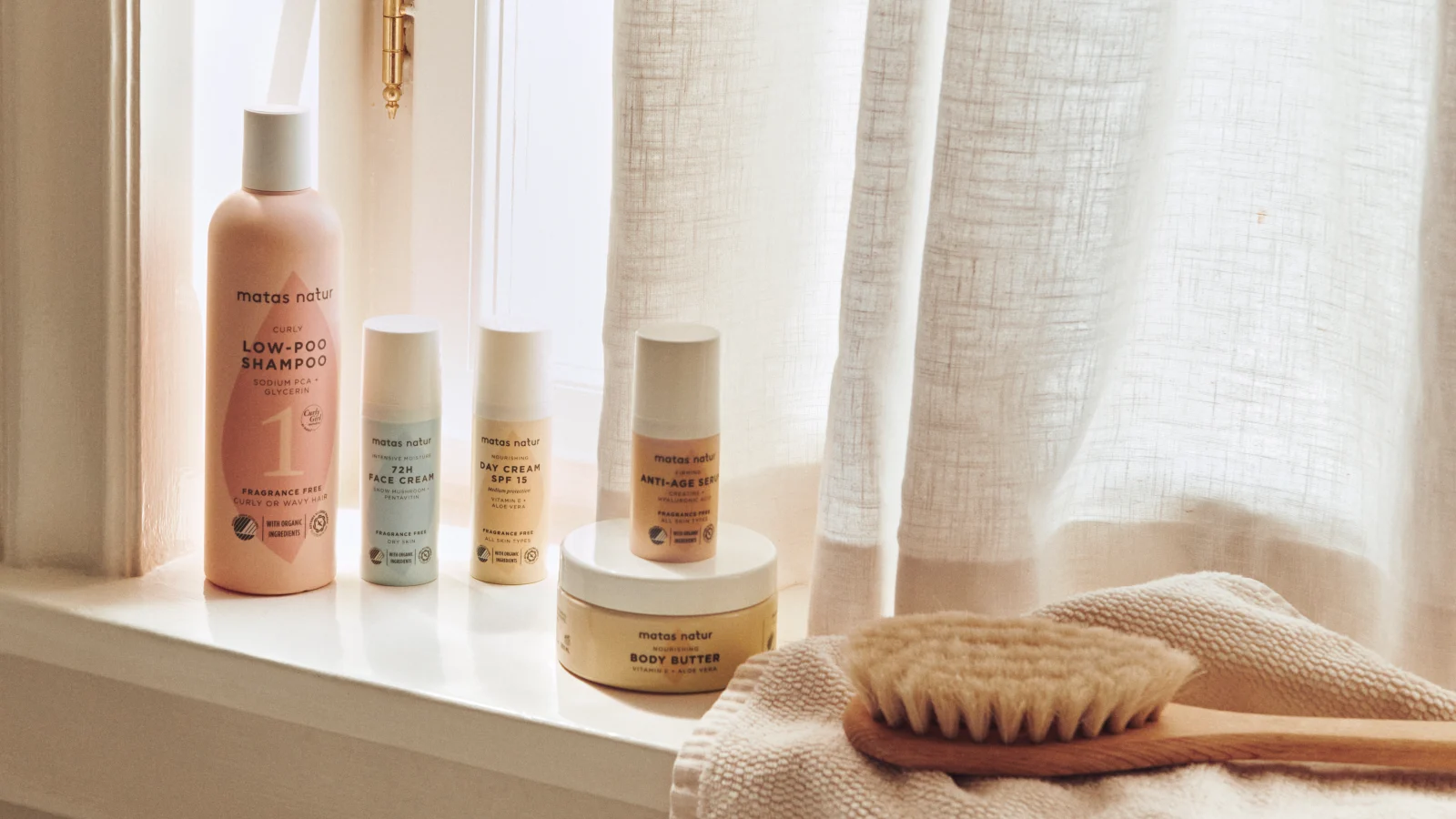 Her ses et stemningsfuldt billede med produkter fra Matas Natur placeret i en lys vindueskarm. Sortimentet omfatter blandt andet Low-Poo Shampoo, ansigtspleje, Day Cream SPF 15, Anti-Age SPF og Body Butter. Ved siden af produkterne ligger en kropsbørste og et tekstil, mens det varme dagslys og de naturlige materialer skaber et roligt og plejende udtryk.