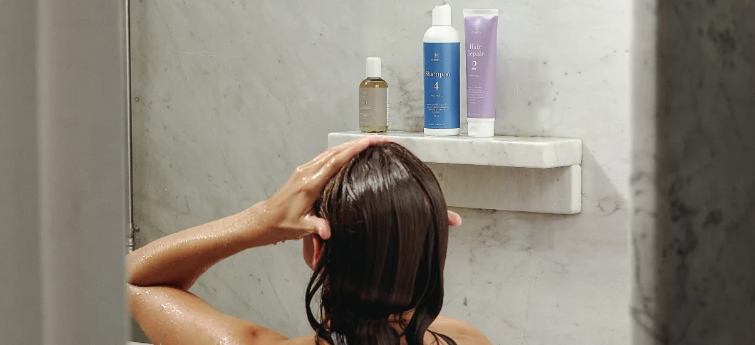 Her ses et billede af en person i badet set bagfra med hårplejeprodukter placeret på en hylde. Produkterne fra Purely Professional inkluderer blandt andet “Shampoo 4” og “Hair Repair 2”.
