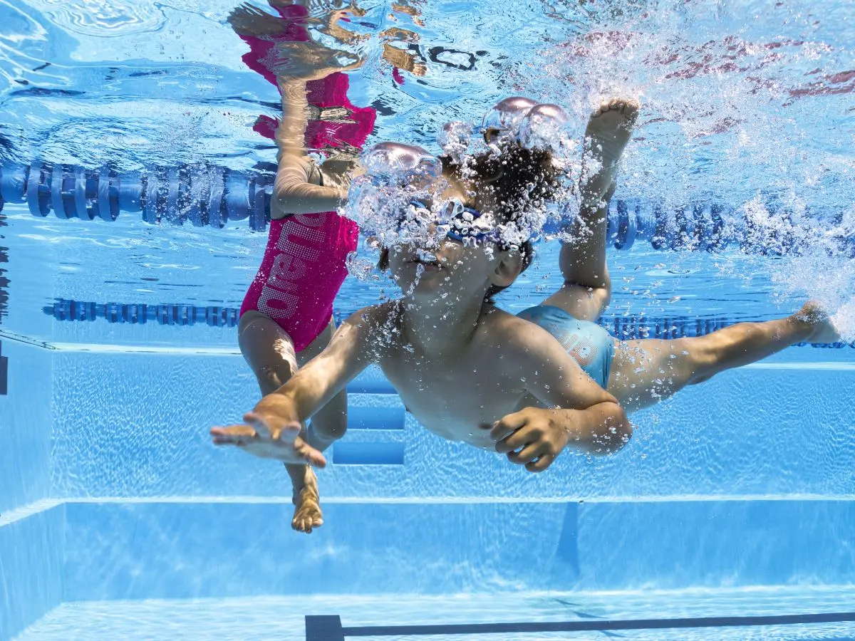 To børn svømmer under vandet i en pool; det ene barn i blå badebukser, det andet i en pink badedragt med teksten 'Varend'.