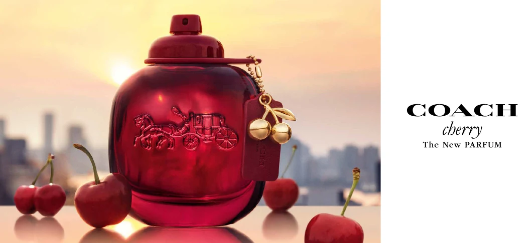 Her ses et billede af en parfumeflakon fra Coach placeret foran en byudsigt ved solnedgang. På billedet står teksten “COACH cherry The New PARFUM”. Flakonen er i mørkerøde nuancer og dekoreret med et kirsebærvedhæng, mens kirsebær er placeret omkring produktet.